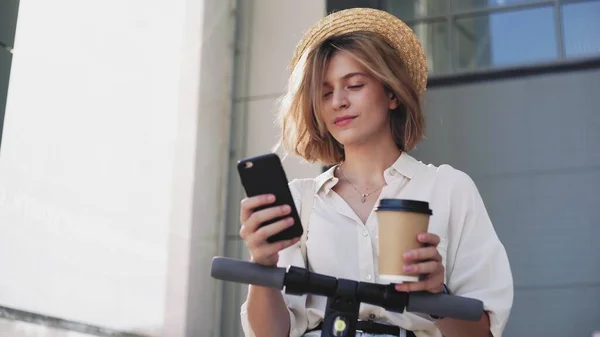 Woman is using smartphone and drinking coffee after ride on electric kick scooter — Stock Photo, Image