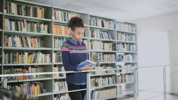 Retrato da jovem afro-americana encaracolada lendo o livro antigo enquanto estava perto das prateleiras de livros na biblioteca . — Fotografia de Stock