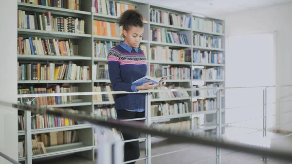 Retrato de la joven afroamericana rizada leyendo el viejo libro mientras estaba de pie cerca de los estantes de los libros en la biblioteca . —  Fotos de Stock