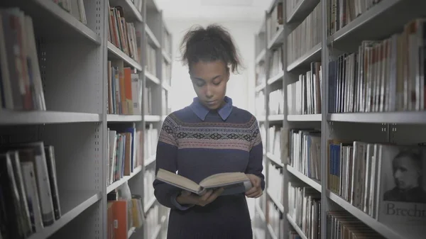 Joven mujer afroamericana rizada está leyendo un libro mientras camina entre las estanterías de la biblioteca . —  Fotos de Stock