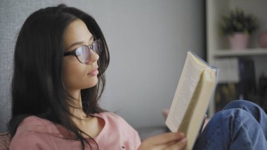 Oldukça güzel, mutlu, gözlüklü bir bayan öğrenci modern bir evin kanepesinde kitap okuyor..