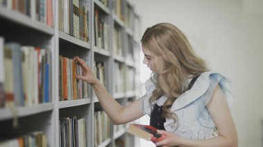 Üniversite kütüphanesindeki raftan kitap alan genç sarışın kadın..