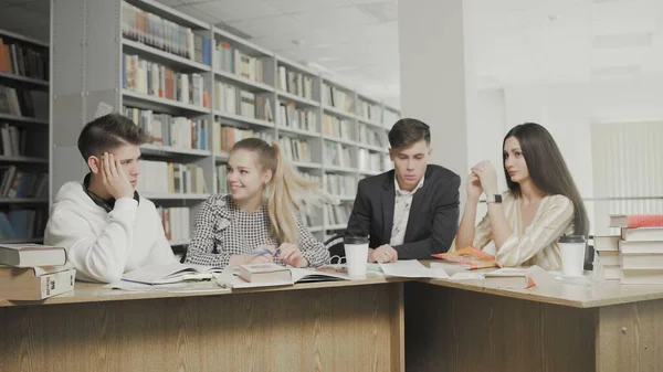 Két férfi és két nő főiskolai hallgató nevet, miközben vizsgákra készülnek, miközben az egyetemi könyvtárban ülnek az asztalnál. — Stock Fotó