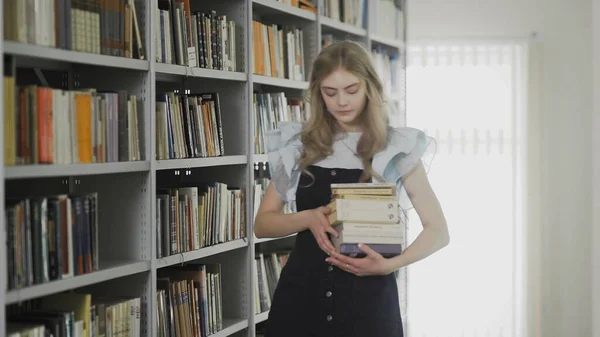 Beautiful young blonde student woman walking in university library with many books in hands — Stock Photo, Image