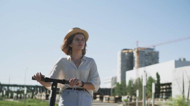 Hasır şapkalı genç bir kadın, elektrikli scooterıyla yürüyor.