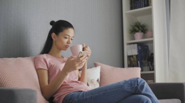 Güzel genç bir kadın akıllı telefon kullanıyor. Kanepede dinleniyor. İnternetten mesaj atıyor, kahve içiyor, rahat bir hayat sürüyor.