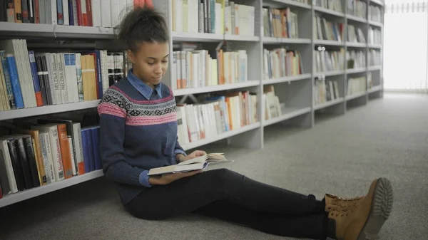 Young african american woman is reading book sitting on floor in university library. — Stock Photo, Image