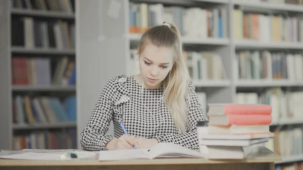 Retrato de una estudiante con auriculares preparándose para el examen y escribiendo notas mientras está sentada en la mesa de la biblioteca universitaria —  Fotos de Stock