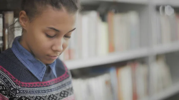 Jovem afro-americana está lendo livro sentado no chão na biblioteca da universidade . — Fotografia de Stock