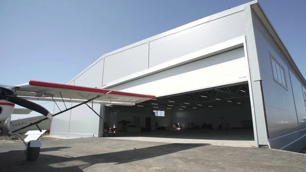 Hangarın dışında küçük uçaklar. Kapı açılıyor. Küçük bir uçağın görünür bir parçası.