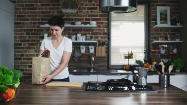 Ev hanımı elinde alışveriş çantasıyla beyaz mutfağa geliyor.