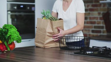 Mutlu genç bayan mutfağa içinde meyve olan bir alışveriş çantası getirir ve paketleri açar..
