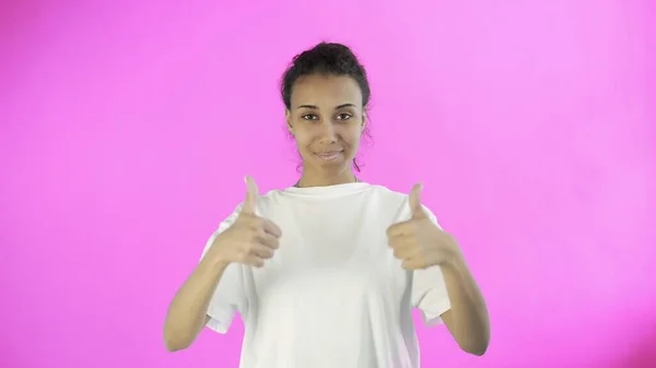 Beautiful young girl in a white T-shirt shows thumb UP with both hands on pink background — Stock Photo, Image