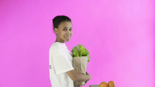 Gelukkig vrouw laden papieren zak met biologische gezonde groenten en fruit op roze achtergrond — Stockfoto