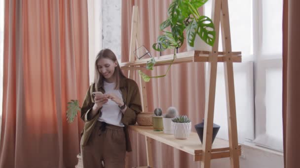 Aantrekkelijke vrouw gebruikt telefoon in de buurt van de plank met bloemen — Stockvideo