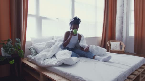 Η όμορφη νεαρή Αφρο-Αμερικανίδα κάθεται στο κρεβάτι, πίνει πρωινό καφέ και χρησιμοποιεί τηλέφωνο. — Αρχείο Βίντεο