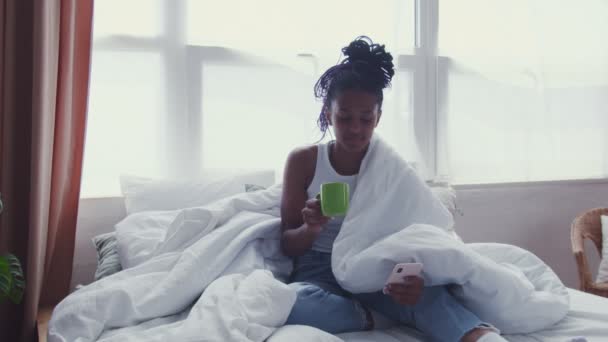Ελκυστική νεαρή Αφροαμερικανή γυναίκα κάθεται στο κρεβάτι, πίνει πρωινό καφέ και χρησιμοποιεί το τηλέφωνο — Αρχείο Βίντεο