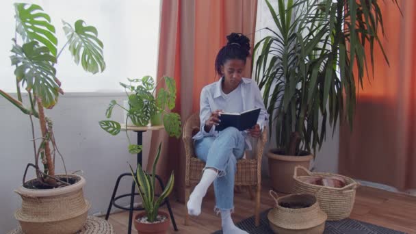 Mujer bastante afroamericana se sienta rodeada de flores en silla de mimbre y lee un libro — Vídeo de stock