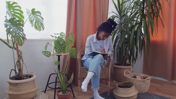 Atractiva joven africana concentrada se sienta rodeada de flores en una silla de mimbre y hace algunas notas en su cuaderno de notas — Vídeo de stock