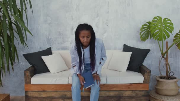 Молода афроамериканка сидить на дивані і читає книжку. — стокове відео