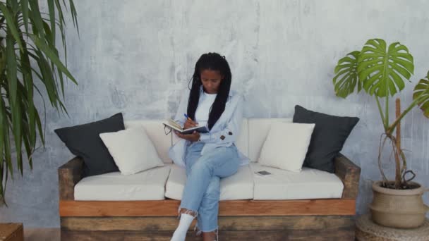 Приваблива зайнята африканка сидить на дивані, розмовляє по телефону і робить деякі нотатки у своїй записниці — стокове відео