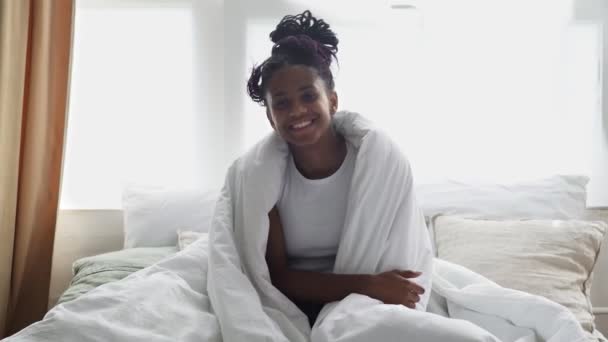 Joven mujer africana feliz se sienta om cama, mira a la cámara y sonríe — Vídeo de stock