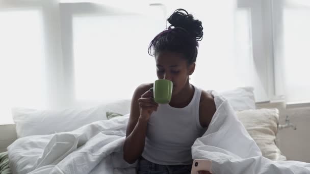 Κοντινό πλάνο του Νεαρή Αφροαμερικανή γυναίκα κάθεται στο κρεβάτι, πίνει πρωινό καφέ και χρησιμοποιεί το τηλέφωνο — Αρχείο Βίντεο
