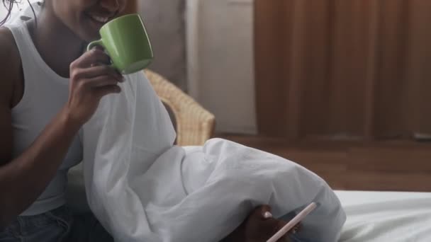 Vista laterale della donna afroamericana si siede a letto, beve caffè del mattino e utilizza il telefono — Video Stock