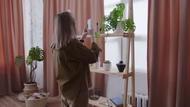 Молодая женщина делает фотографии своих домашних растений на полке — стоковое видео