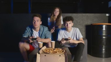 Oyun konsolunda iki erkek ve bir kız oynuyor. Koltukta oturuyorlar ve joystickleri tutuyorlar. Bir adam diğerini yeniyor.