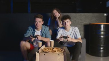 Arkadaşlar oyun konsolunda oynar. Koltukta oturuyorlar ve joystickleri tutuyorlar. Bir adam diğerini yeniyor.