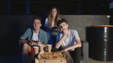 Bir grup arkadaş karanlık odada oyun konsolunda oynarken eğleniyorlar. Bir adam pizza yiyor.