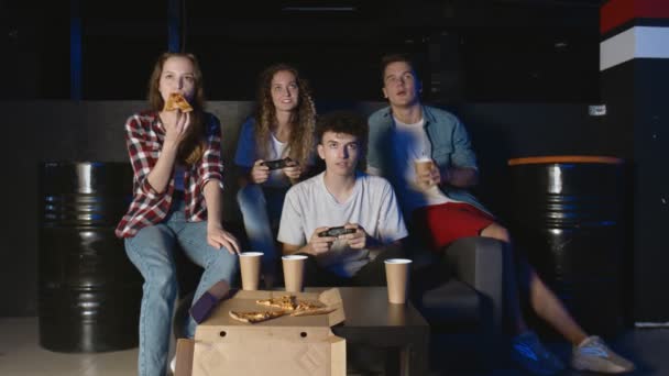 Jovens homens e mulheres alegres estão se divertindo com o videogame na festa em casa enquanto os amigos riem assistindo, conversando e comendo pizza — Vídeo de Stock
