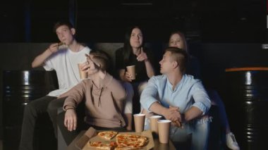 Genç arkadaşlar futbol izleyip pizza yemekten ve karanlık odada kanepede oturmaktan zevk alıyorlar.
