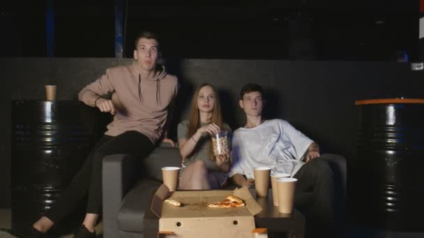 Gli amici si siedono sul divano, guardano film divertenti in TV. Giovane gruppo caucasico seduto sul divano, bevande e spuntini. Serata film. — Video Stock