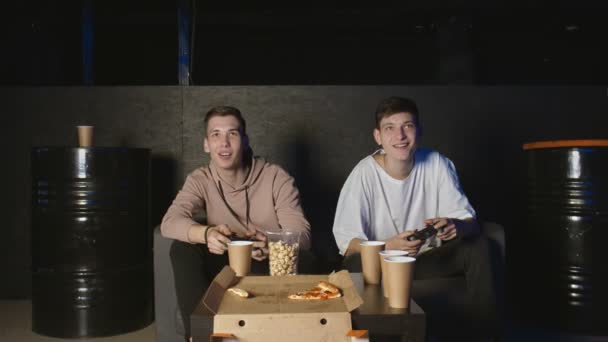 两个年轻人在黑暗的房间里玩电子游戏，坐在沙发上 — 图库视频影像