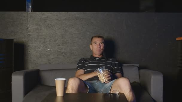 Homme adulte assis sur le canapé, mange du pop-corn et regarde la télévision — Video