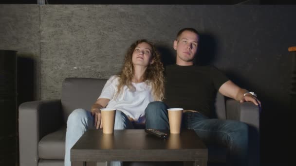 Krásná dívka a mladý muž sedí na gauči a dívá se na televizi. Lidé odpočívají na gauči. — Stock video
