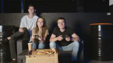 Arkadaşlar oyun konsolunda oynar. Koltukta oturuyorlar ve joystickleri tutuyorlar. Bir adam diğerini yeniyor.