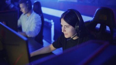 Güzel bir kadın modern bir bilgisayar kulübünde online oyun oynuyor. Genç kadın kulaklıkla monitörün önünde oturuyor, dikkatli bakıyor ve gülüyor..