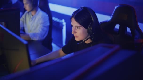 Νεαρή γυναίκα κάθεται σε ακουστικά σε μια σύγχρονη λέσχη υπολογιστών, παίζει ένα online παιχνίδι. Γυναίκα χαμογελά και είναι έκπληκτος με το παιχνίδι στο διαδίκτυο. Προβολή πορτραίτου. — Αρχείο Βίντεο