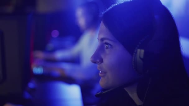 Hermosa mujer juega un juego en línea en un moderno club de informática. Mujer joven se sienta en los auriculares delante del monitor, mira con atención y se ríe. Emociones de alegría y sorpresa en un juego de ordenador — Vídeo de stock