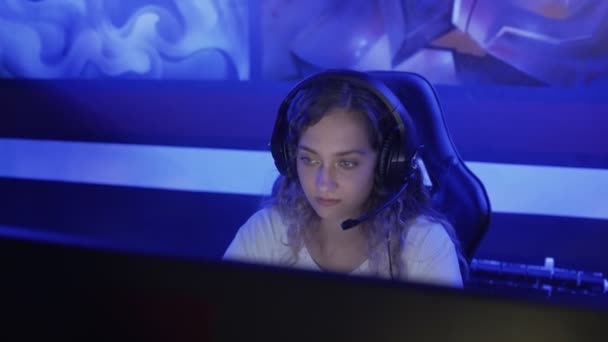 Mujer joven se sienta en los auriculares en un club de informática moderna, juega un juego en línea. Mujer sonríe y se sorprende con el juego de Internet. Vista de retrato. — Vídeos de Stock