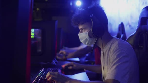 Vista lateral del hombre joven en la máscara de la cara juega en el videojuego en línea en el club de computadoras moderno. — Vídeo de stock