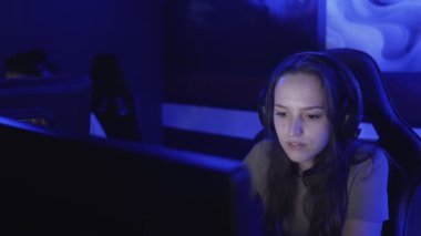 Genç bir kadın modern bir bilgisayar kulübünde kulaklıkla oturup online oyun oynuyor. Kadın gülümsüyor ve internet oyununa şaşırıyor. Dikey görünüm.