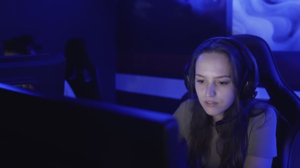 Egy fiatal nő fülhallgatóban ül egy modern számítógépes klubban, online játékot játszik. A nő mosolyog, és meglepődik az internetes játék. Portré nézet. — Stock videók