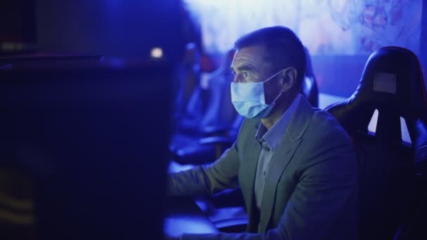 Vue latérale du vieil homme en costume et masque facial bavarder et surfer sur Internet dans le club informatique moderne. — Video