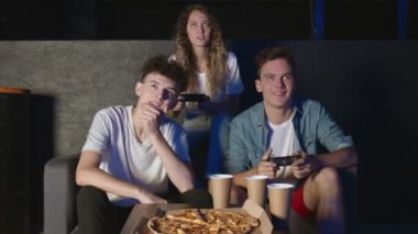 Bir grup arkadaş karanlık odada oyun konsolunda oynarken eğleniyorlar. Bir adam pizza yiyor.