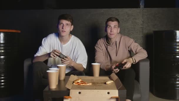 Два счастливых друга играют в экшн-видеоигру в гостиной, сидя на диване. Интенсивная конкуренция между двумя молодыми игроками мужского пола полна эмоций. — стоковое видео