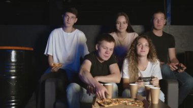 Bir grup arkadaş oturma odasında kanepede dinleniyor, video oyunları oynuyor ve pizza yiyorlar.